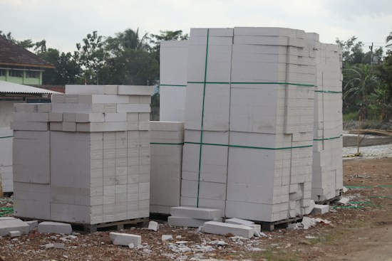Large stacks of white concrete blocks are neatly arranged on wooden pallets. They are outside, beside a dirt path with some debris scattered around. Trees and a building with a corrugated metal roof are visible in the background.