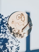 A round beige surface holds several cosmetic and jewelry items. There is a tube of skincare cream, a pair of golden earrings, a necklace featuring pearl elements, and a few transparent capsules. The surface is lightly shadowed with leaf patterns, suggesting the presence of plants nearby.