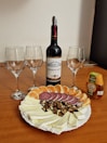 A bottle of Bordeaux wine is placed centrally on a wooden table, surrounded by three empty wine glasses. In front of the bottle is a platter with slices of soft cheese, salami, walnut pieces, and mandarin orange segments. A small bottle of acacia honey is positioned to the right of the wine bottle.