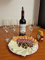 A bottle of Bordeaux wine is placed centrally on a wooden table, surrounded by three empty wine glasses. In front of the bottle is a platter with slices of soft cheese, salami, walnut pieces, and mandarin orange segments. A small bottle of acacia honey is positioned to the right of the wine bottle.