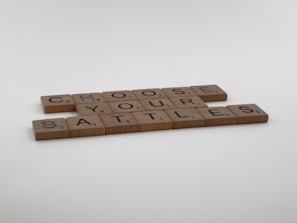 Wooden tiles arranged to spell 'Choose Your Battles', resembling Scrabble pieces on a plain light gray background.