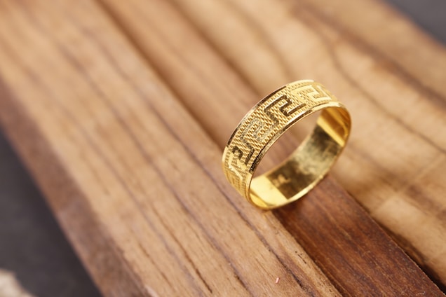 A gold ring with intricate geometric patterns rests elegantly on a wooden surface. The ring's texture and design catches the light, drawing attention to its craftsmanship.