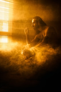 A person sits contemplatively in a hazy, warmly lit room with golden smoke surrounding them. The individual holds a mask, partially hidden by the smoke and light streaming through window blinds.