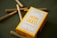 A yellow box labeled 'MOM GRASS' containing five pre-rolled 100% organic hemp flower joints is displayed on a dark surface. Several pre-rolled joints are scattered around the box.