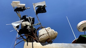 Fabric and plastic flags are attached to a structure made of irregularly shaped concrete and metal rods. A clear blue sky serves as the backdrop, and part of a boat's equipment, possibly radar, is visible.