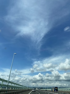 A highway stretches into the distance under a bright blue sky filled with fluffy clouds. Cars are traveling along the road, and tall streetlights line the highway. The bridge structure and railings are visible alongside the road.