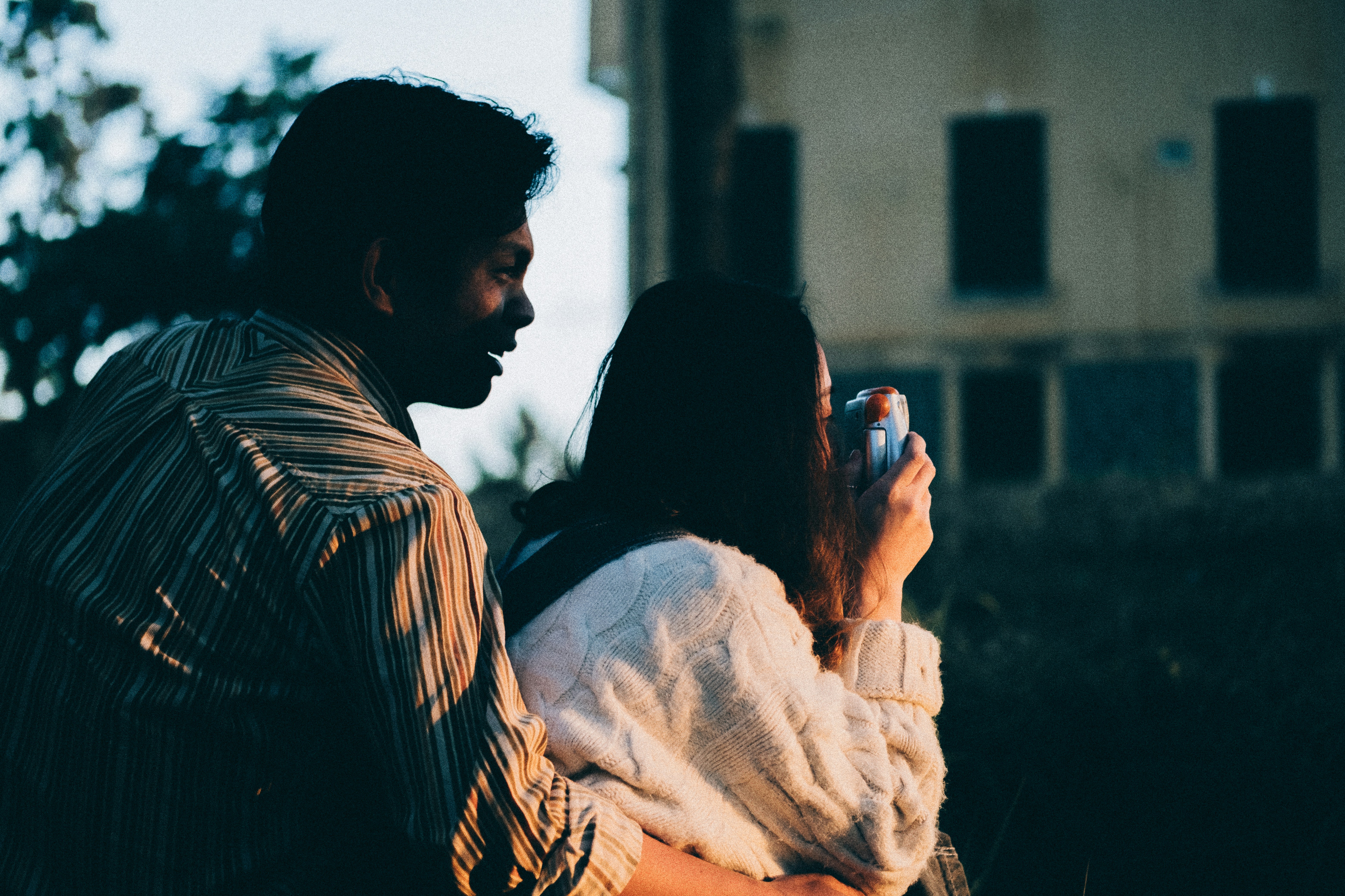 Videographer filming couple at sunset, professional cinema camera on shoulder, dramatic backlight silhouette, warm dusk sky