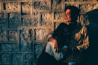 A couple embraces against a textured stone wall, bathed in warm, golden light. The person on the right has a contemplative expression, looking into the distance, while the other leans in closely, partly in shadow.
