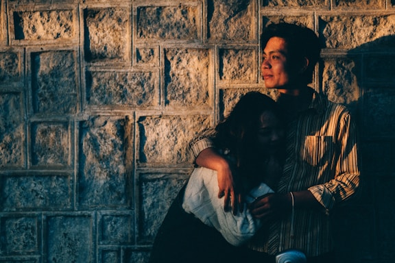 A couple embraces against a textured stone wall, bathed in warm, golden light. The person on the right has a contemplative expression, looking into the distance, while the other leans in closely, partly in shadow.