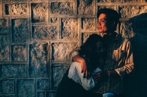 A couple embraces against a textured stone wall, bathed in warm, golden light. The person on the right has a contemplative expression, looking into the distance, while the other leans in closely, partly in shadow.