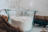 On a kitchen counter, a clear glass measuring cup is filled with white flour. Nearby, there is a spool of brown twine, and the background is softly blurred.
