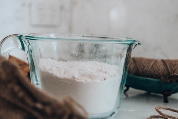 On a kitchen counter, a clear glass measuring cup is filled with white flour. Nearby, there is a spool of brown twine, and the background is softly blurred.