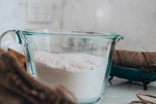On a kitchen counter, a clear glass measuring cup is filled with white flour. Nearby, there is a spool of brown twine, and the background is softly blurred.