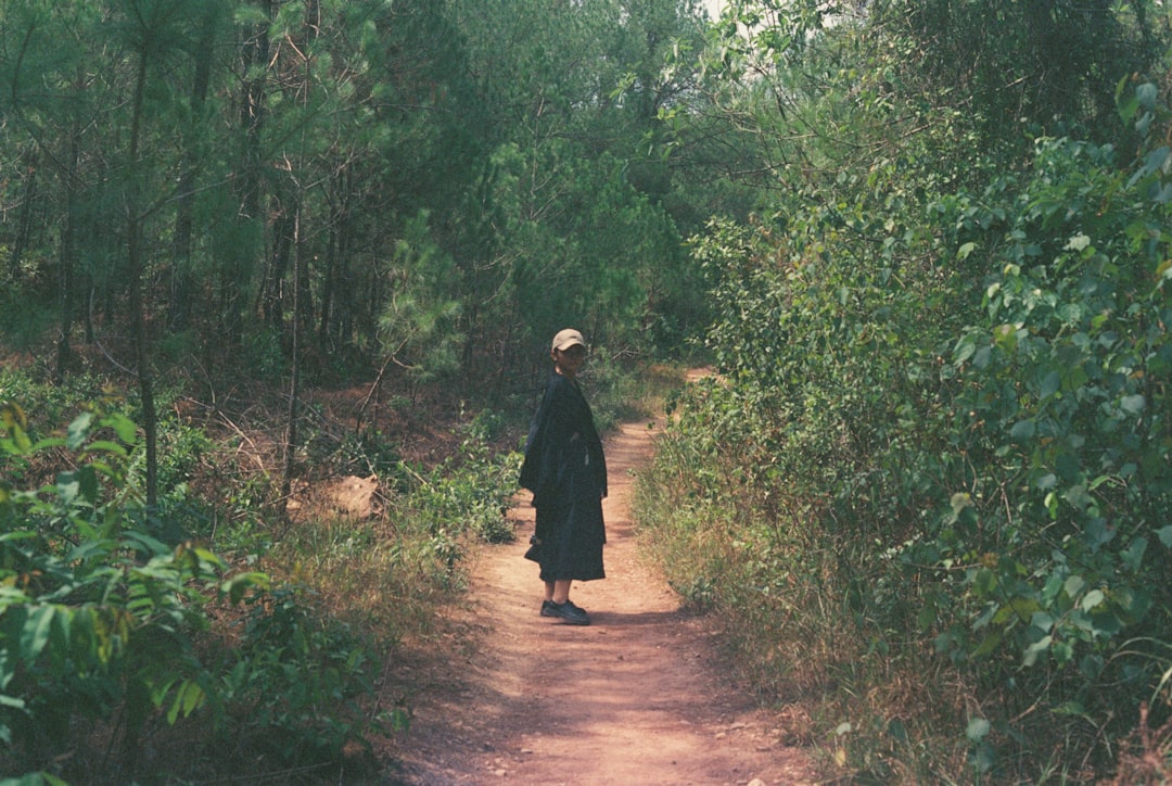 woman in black coat walking on dirt road between green trees during daytime