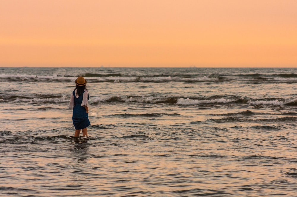 homem de camisa azul e calção preto em pé na costa do mar durante o pôr do sol