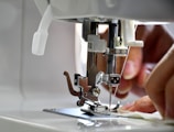 A close-up view of a sewing machine with hands guiding fabric under the presser foot. The needle is prominently in focus, indicating an active sewing process.