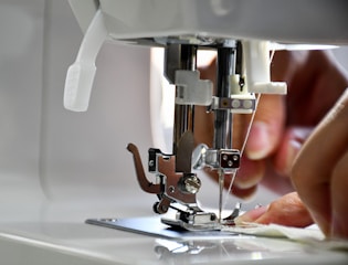 A close-up view of a sewing machine with hands guiding fabric under the presser foot. The needle is prominently in focus, indicating an active sewing process.