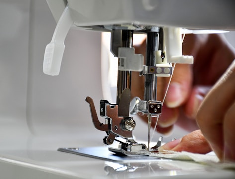 A close-up view of a sewing machine with hands guiding fabric under the presser foot. The needle is prominently in focus, indicating an active sewing process.