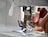 A close-up view of a sewing machine with hands guiding fabric under the presser foot. The needle is prominently in focus, indicating an active sewing process.