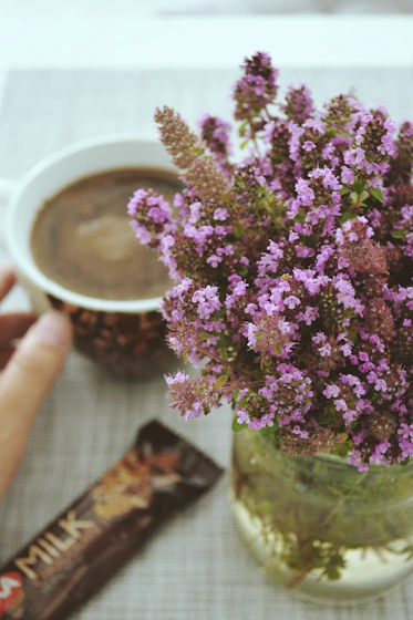Close-up of hands gently holding a steaming cup of cacao with purple flowers around.