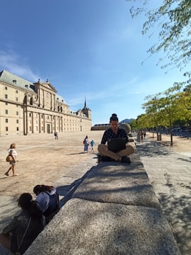 A person is sitting cross-legged on a stone wall, working on a laptop in an open plaza with a large historic building in the background. The sky is clear blue, and several people are walking or sitting nearby. Trees line the path to the right, casting shadows on the ground.