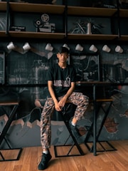 A young man is sitting on a stool in a stylish cafe with a wall decorated with artistic elements like a film reel and a turntable. He is wearing a black T-shirt with the word 'Jordan' and camouflage-patterned pants. Above him are white coffee mugs hanging on a grid.