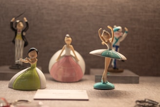 A collection of whimsical figurines displayed on a shelf, featuring characters such as a ballerina in a white and green dress, a flutist in a green and white outfit, and a conductor in the background. The scene has a warm, soft lighting that highlights the playful and artistic nature of the figures.