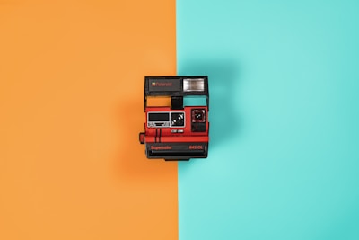 A vintage instant camera is centered against a background split into two vibrant colors: orange on the left and teal on the right. The camera is predominantly red and black with a flash unit on top and branding that includes 'Supercolor 645 CL.'