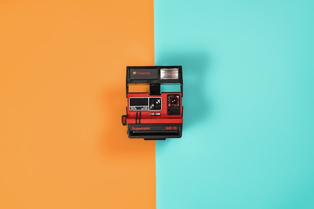 A vintage instant camera is centered against a background split into two vibrant colors: orange on the left and teal on the right. The camera is predominantly red and black with a flash unit on top and branding that includes 'Supercolor 645 CL.'