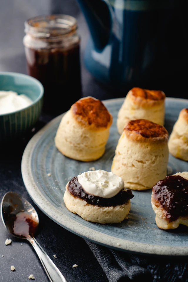 Scones on a blue stoneware plate, one topped with jam and whipped cream