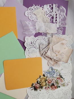 Various craft materials are arranged, including lace fabric, doilies, pastel paper cards, aged text paper squares, and floral cutouts. The lace and doilies display intricate patterns, the paper is pale and weathered, and the floral designs feature vibrant colors against a soft backdrop.