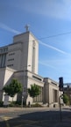 A large, imposing modern building with light-colored stone walls and a tall tower featuring an antenna on top. The building is surrounded by a few trees and is located next to a street with a visible traffic light. The sky is clear with a few contrails visible.