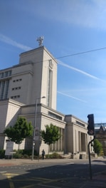 A large, imposing modern building with light-colored stone walls and a tall tower featuring an antenna on top. The building is surrounded by a few trees and is located next to a street with a visible traffic light. The sky is clear with a few contrails visible.