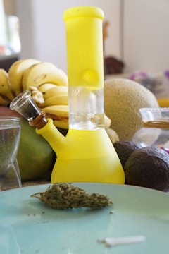 A yellow glass bong is placed on a table surrounded by various fruits such as bananas, a melon, and avocados. A glass of water and a nugget of cannabis are also on the table. The scene is set in a kitchen or dining area with a casual atmosphere.