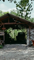 A rustic structure with a sloped roof, built from wooden beams and supported by bags filled with material, stands amidst a lush, green landscape. Inside, a vehicle is partially visible in the shadowed interior. Surrounding vegetation includes tall trees and dense foliage, offering a sense of seclusion and tranquility.