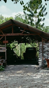 A rustic structure with a sloped roof, built from wooden beams and supported by bags filled with material, stands amidst a lush, green landscape. Inside, a vehicle is partially visible in the shadowed interior. Surrounding vegetation includes tall trees and dense foliage, offering a sense of seclusion and tranquility.