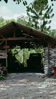 A rustic structure with a sloped roof, built from wooden beams and supported by bags filled with material, stands amidst a lush, green landscape. Inside, a vehicle is partially visible in the shadowed interior. Surrounding vegetation includes tall trees and dense foliage, offering a sense of seclusion and tranquility.