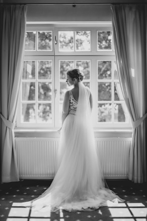A bride stands gracefully in front of a large window, the natural light illuminating her white wedding dress. The setting has an elegant and serene atmosphere, with curtains drawn to the sides and a patterned carpet beneath her feet.