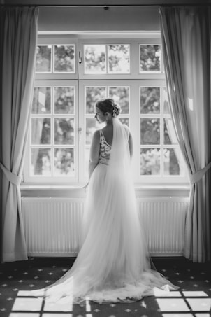 A bride stands gracefully in front of a large window, the natural light illuminating her white wedding dress. The setting has an elegant and serene atmosphere, with curtains drawn to the sides and a patterned carpet beneath her feet.