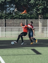 Two soccer players are engaged in action on a soccer field. One player wearing a red jersey is controlling the ball while being closely challenged by another player in a navy-blue jersey. The field is green with patches of artificial turf and there is a metal fence in the background with some trees and greenery.
