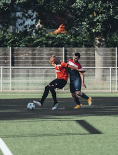 Two soccer players are engaged in action on a soccer field. One player wearing a red jersey is controlling the ball while being closely challenged by another player in a navy-blue jersey. The field is green with patches of artificial turf and there is a metal fence in the background with some trees and greenery.
