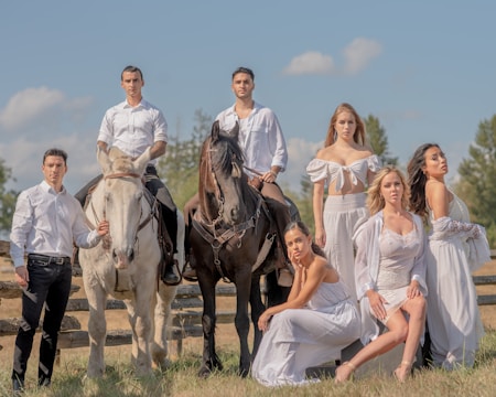 A group of people are posing outdoors with two horses, all dressed in white attire. The setting is a sunny, open field with a wooden fence and trees in the background. Two men are seated on the horses, while the others stand or kneel beside them, radiating a sense of elegance and unity.