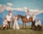 A group of people stands and sits on a grass field in front of a majestic mountain range under a blue sky with clouds. Two horses, one brown and one white, are present, with one person sitting on the brown horse. The group is dressed in coordinated light or white outfits, giving a harmonious and elegant look.