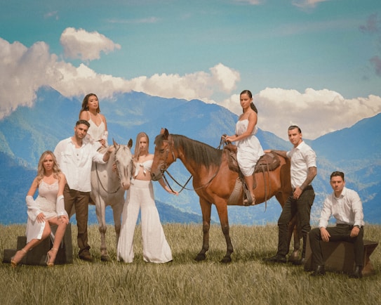 A group of people stands and sits on a grass field in front of a majestic mountain range under a blue sky with clouds. Two horses, one brown and one white, are present, with one person sitting on the brown horse. The group is dressed in coordinated light or white outfits, giving a harmonious and elegant look.