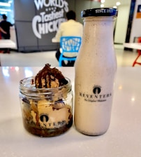 A glass bottle and a jar of dessert are placed on a table. The bottle has a label reading 'Keventers' and contains a creamy beverage. The jar is filled with ice cream topped with a chocolate drizzle. In the background, there are people sitting on chairs and a sign on the wall that reads 'World's Tastiest Chicken.'