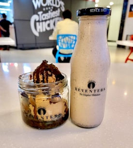 A glass bottle and a jar of dessert are placed on a table. The bottle has a label reading 'Keventers' and contains a creamy beverage. The jar is filled with ice cream topped with a chocolate drizzle. In the background, there are people sitting on chairs and a sign on the wall that reads 'World's Tastiest Chicken.'