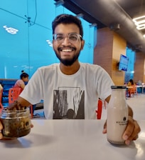 A smiling man with glasses is seated at a table in a well-lit eating area. He is holding a jar with some kind of dessert in his left hand and a bottle of Keventers beverage in his right hand. He is wearing a white T-shirt with an abstract cityscape design. In the background, there are other people sitting at tables and a television screen mounted on the wooden wall.