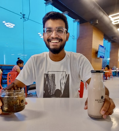 A smiling man with glasses is seated at a table in a well-lit eating area. He is holding a jar with some kind of dessert in his left hand and a bottle of Keventers beverage in his right hand. He is wearing a white T-shirt with an abstract cityscape design. In the background, there are other people sitting at tables and a television screen mounted on the wooden wall.