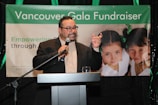 A person in a suit is speaking at a podium holding a microphone, with a banner in the background that reads 'Vancouver Gala Fundraiser'. The banner also features two young girls with green bows in their hair.