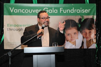 A person in a suit is speaking at a podium holding a microphone, with a banner in the background that reads 'Vancouver Gala Fundraiser'. The banner also features two young girls with green bows in their hair.